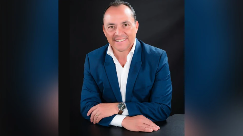 A smiling man with short black hair wearing a navy blue suit jacket, white dress shirt and brown watch on his left wrist rests his crossed arms on a black surface in front of a black background. He's visible from the waist up.