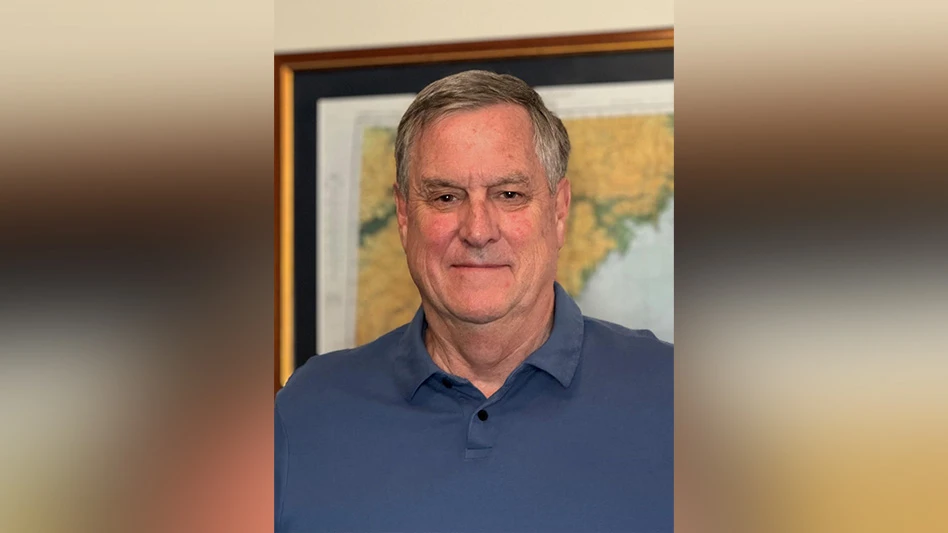 A man with short gray hair wearing a dark blue polo shirt stands in front of a framed map. He is visible from the shoulders up.