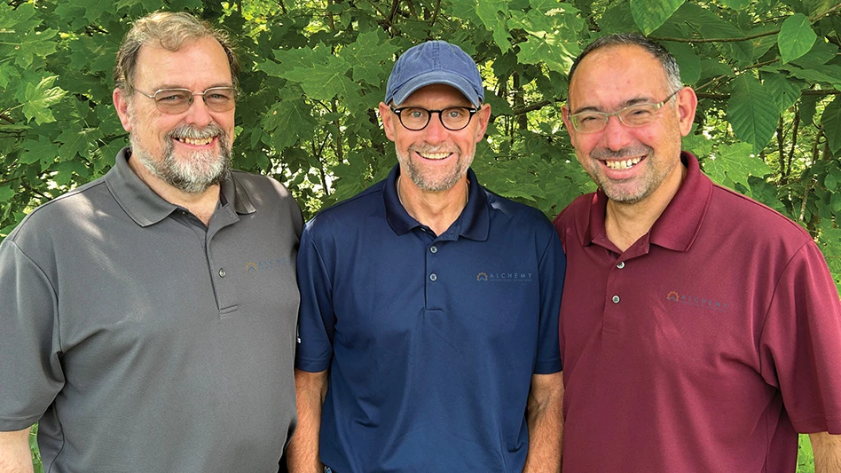 A photo of three smiling men standing in front of green leaves. The man on the left has short gray hair and a beard and is wearing glasses and a gray polo shirt. The man in the middle has a short gray beard and is wearing a blue ball cap, black glasses and a navy blue polo shirt. The man on the right has short black hair and is wearing glasses and a maroon polo shirt.