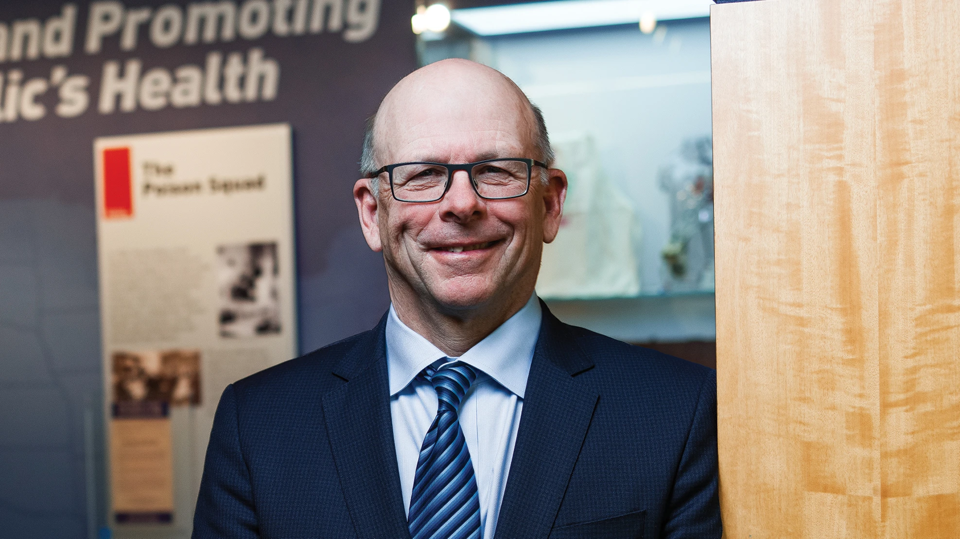 A smiling bald man wearing black glasses, a light blue dress shirt, a navy blue suit jacket and a blue striped tie.