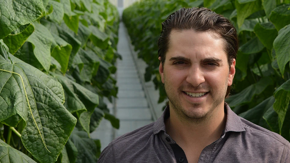 A photo of a smiling man with short brown hair wearing a gray shirt and standing in a greenhouse with a row of leafy greens behind him.