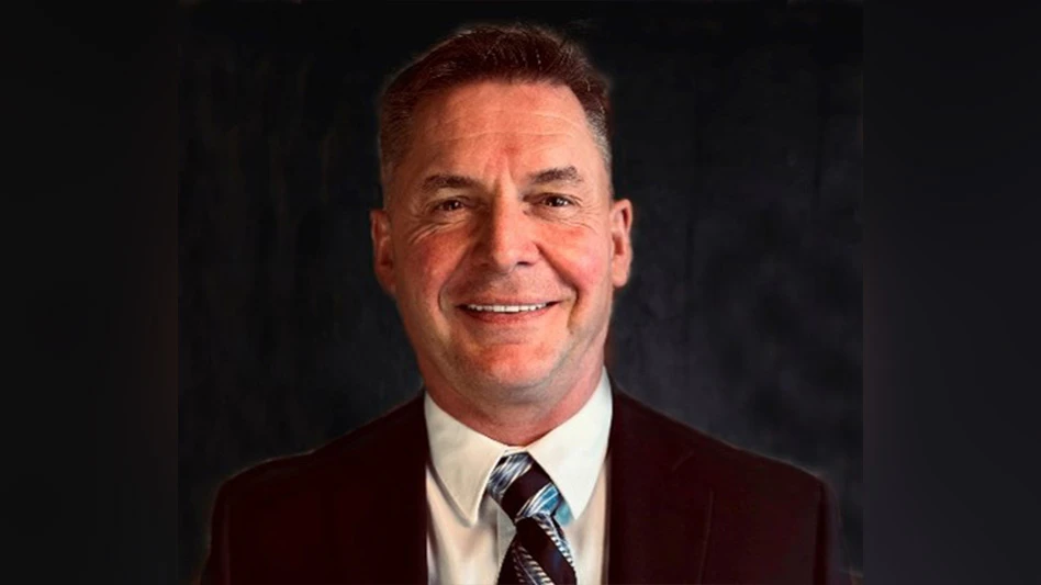 A headshot photo of a smiling man with short brown hair wearing a black suit, white shirt and tie.