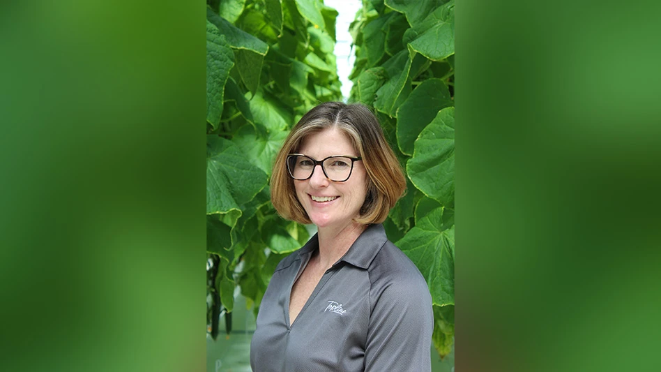 A smiling woman with short blonde hair wearing black glasses and a gray collared shirt stands in front of leafy greens.
