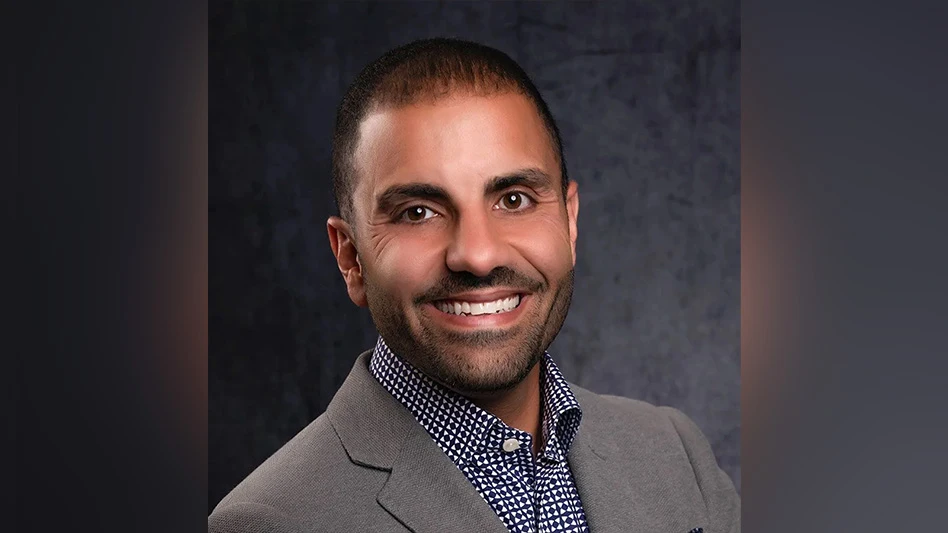 A photo of a smiling man with short black hair and short black facial hair and wearing a gray suit jacket and blue and white plaid button-up shirt.