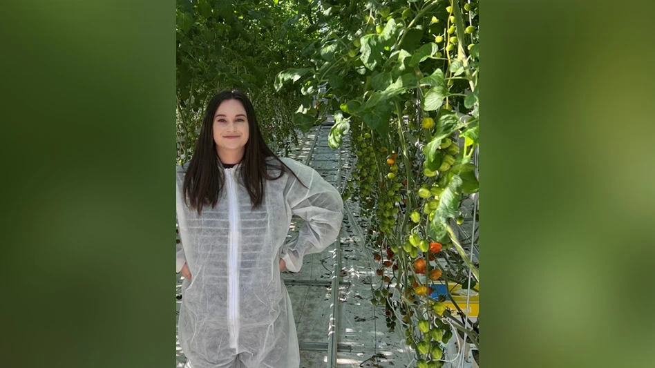 A woman with long brown hair wearing a white protective suit stands with her hands on her hips in a greenhouse surrounded by green and red tomatoes on green vines.
