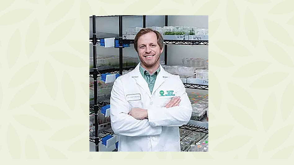 A man in a white lab coat standing, arms crossed, in front of lab shelves.