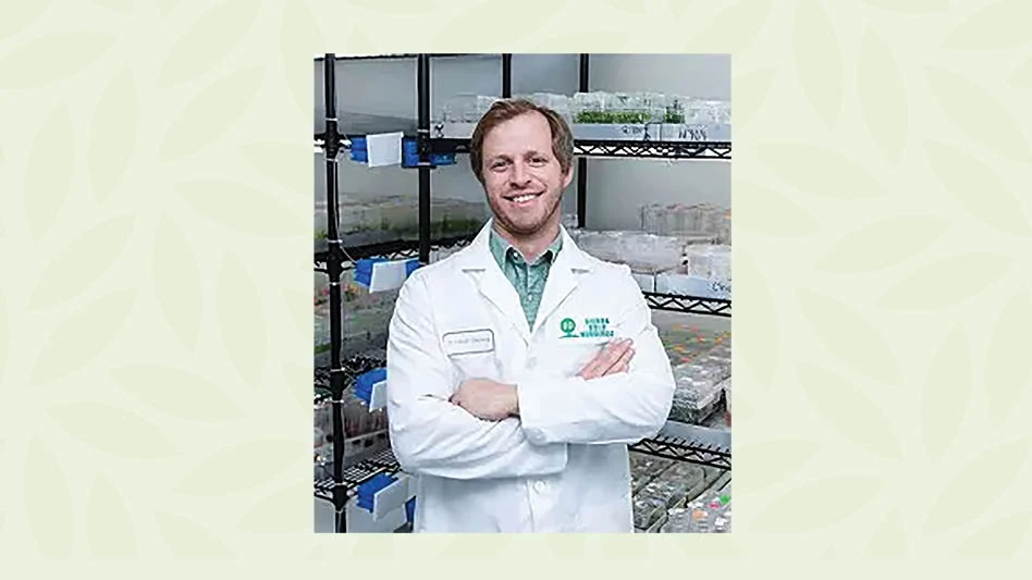 A man in a white lab coat standing, arms crossed, in front of lab shelves.