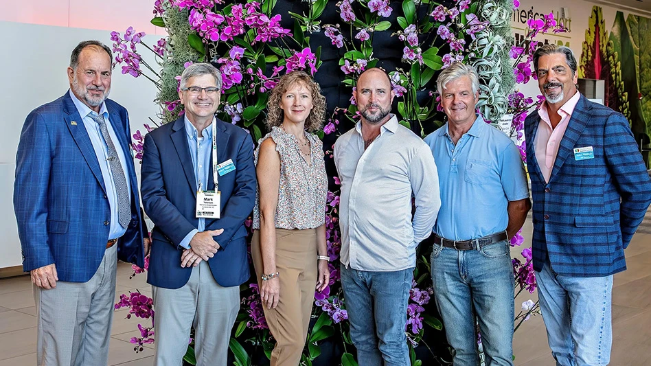 Six smiling people pose in front of a column of pink flowers and green leaves.