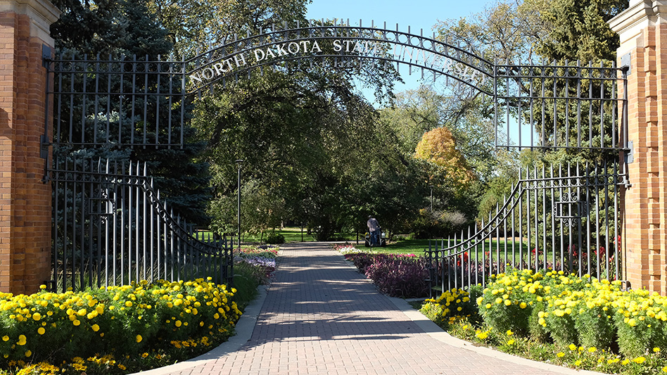 A black metal entrance gate reading North Dakota State University connected to two brick pillars and spanning over a brick path with flowers growing along it.